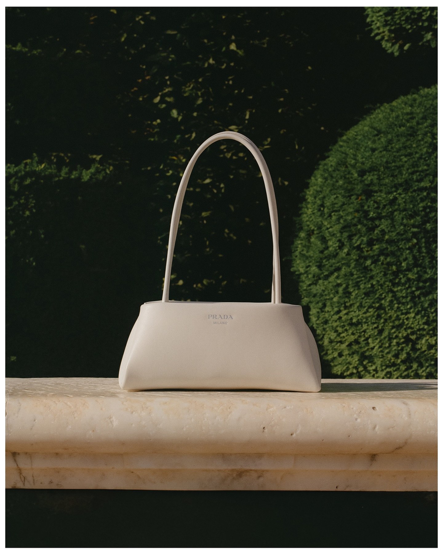 A pale leather handbag on a marble ledge in front of a clipped hedge