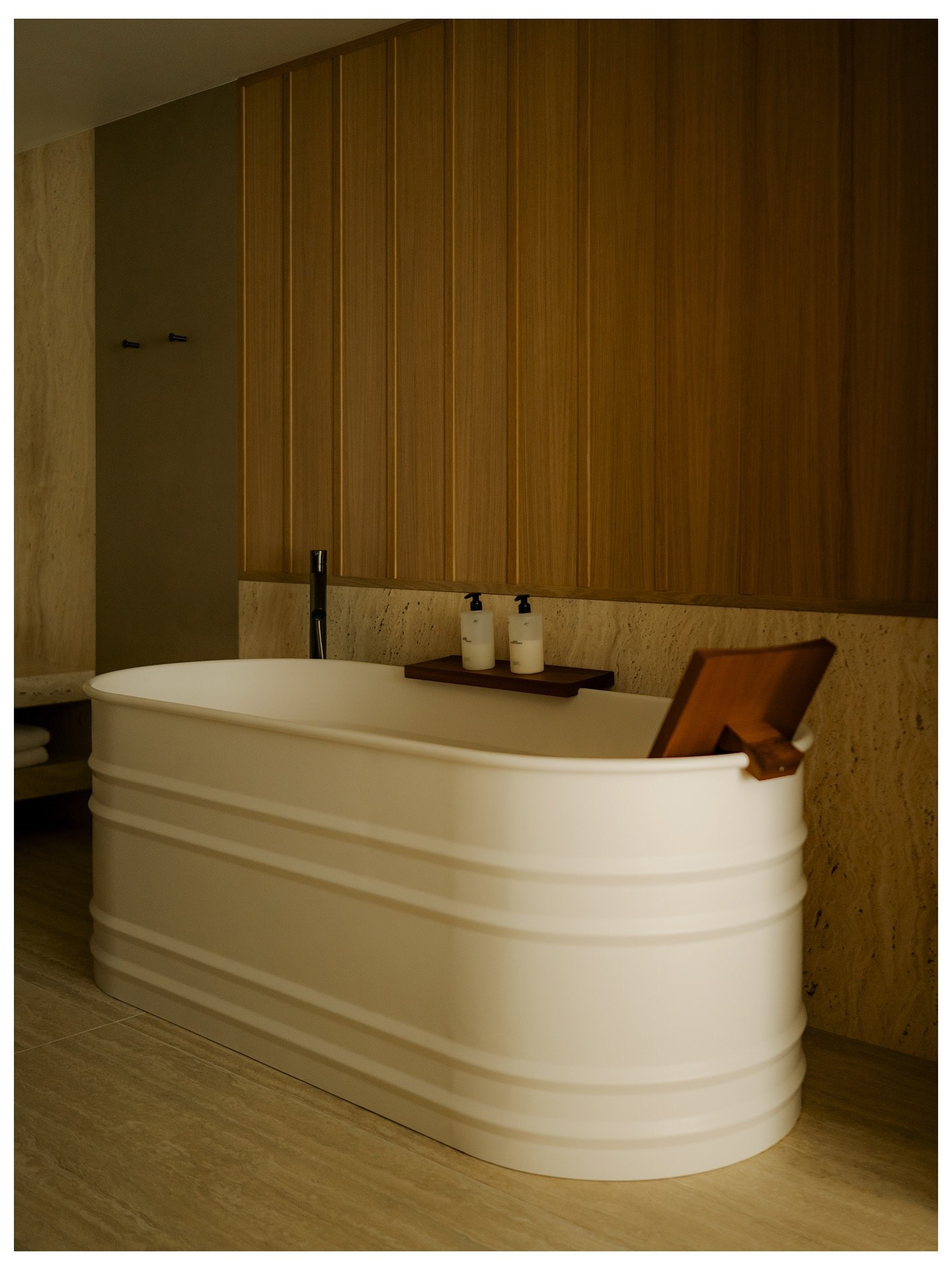 A freestanding bath against travertine and oak panelling in low light